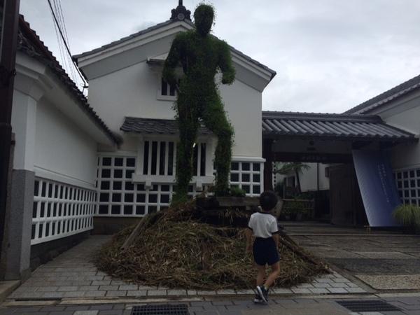 家の高さほどある巨大な緑の人型のオブジェを小さい子供が見上げている写真